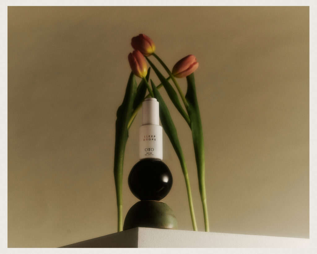 A skincare bottle is displayed on top of two stacked spheres, with two tulips arching over it, all set against a soft, neutral background.
