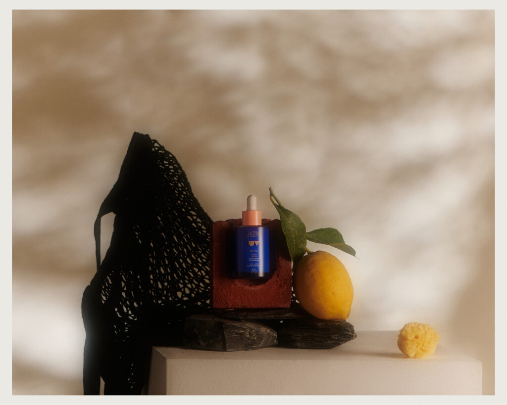 A blue dropper bottle sits on a brick with a lemon, sponge, black netted fabric, and dark stones arranged nearby, set against a softly lit, shadowy background.