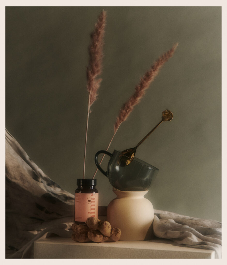 A still life featuring a bottle of supplements, pieces of ginger root, a green glass cup inside a beige vase, a golden stirring stick, and tall dried grasses against a muted background with draped fabric.