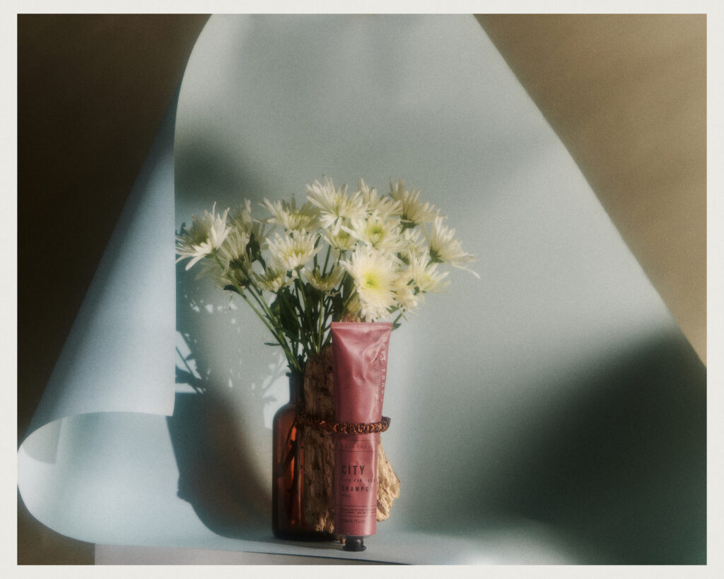 A bouquet of white daisies in a glass vase sits beside a pink tube of cream, with soft light and shadows cast on a pastel blue backdrop.