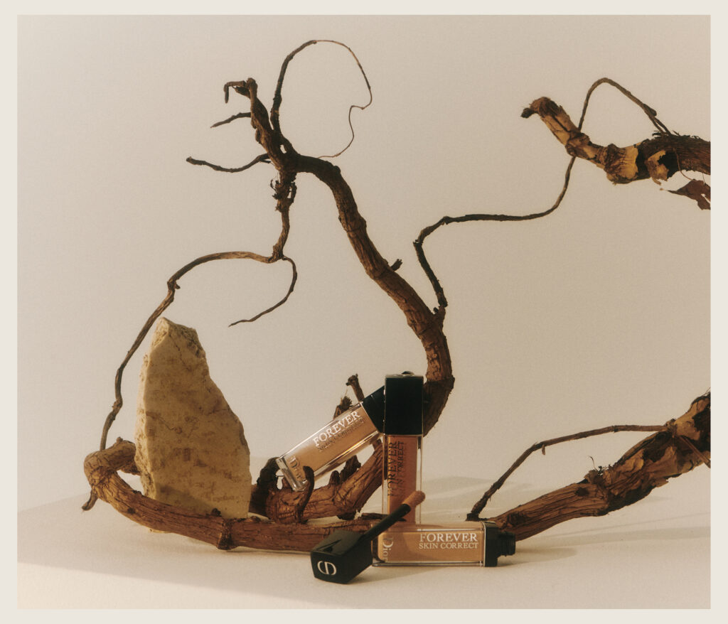 Two tubes of Forever Skin Correct concealer are displayed among dried, twisted branches and a rough stone, set against a beige background in soft, warm lighting.
