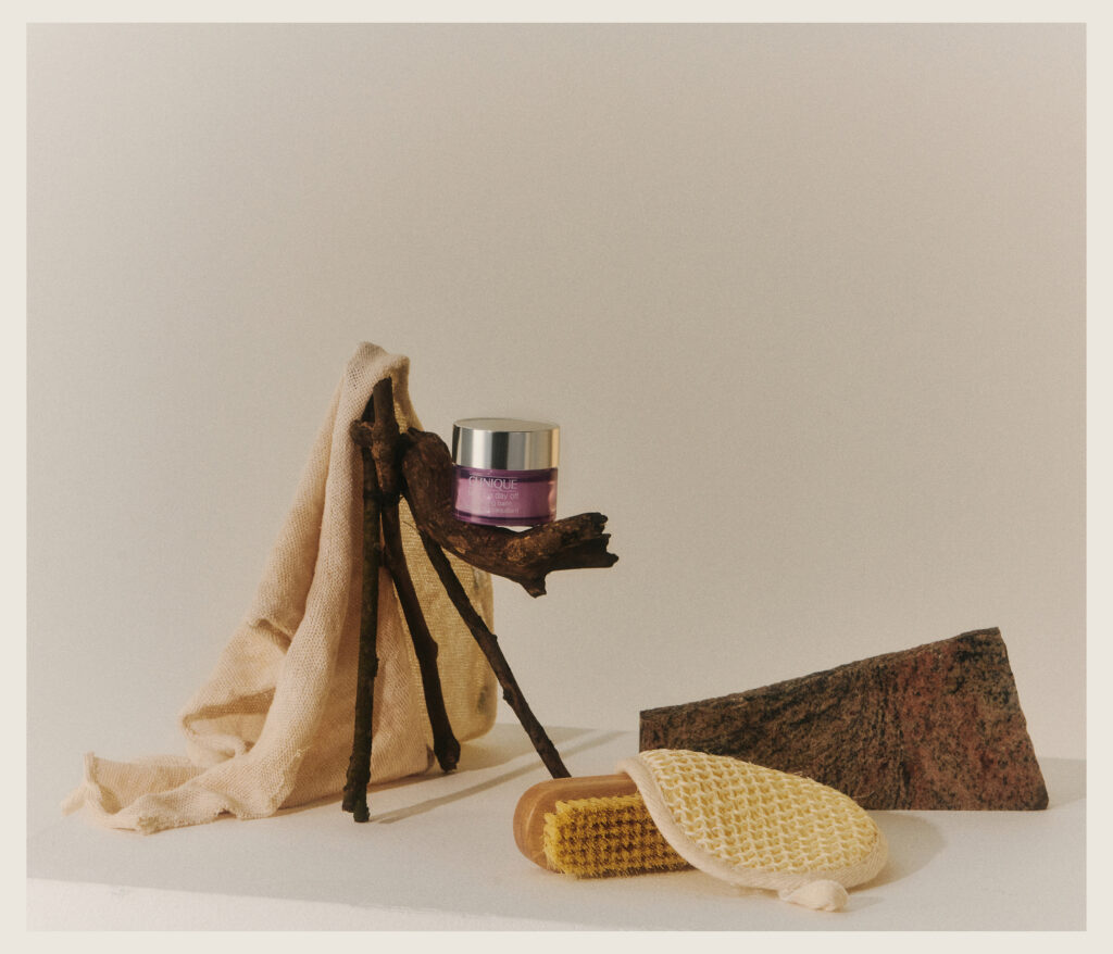 A purple cosmetic jar with a silver lid sits on a small branch stand draped with beige fabric. Nearby are a textured brown stone and a yellow brush with a woven top and a handle. The background is neutral.