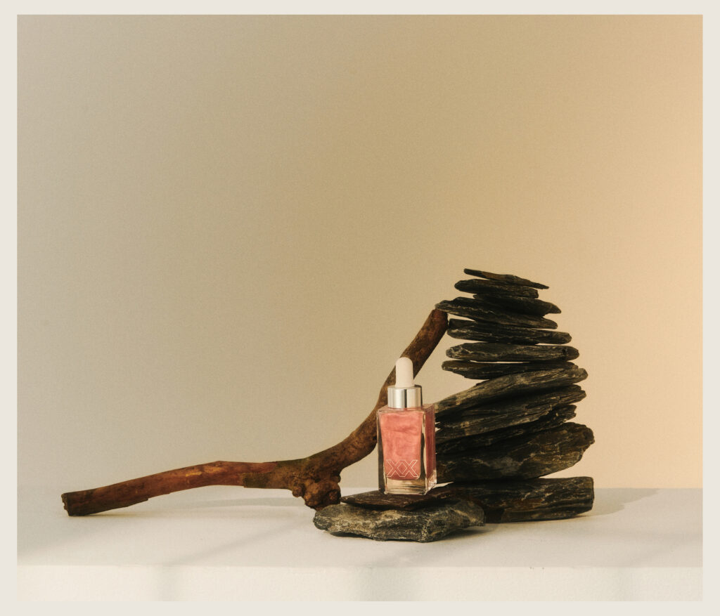 A glass dropper bottle with pink liquid stands on a flat stone, next to a stacked arrangement of dark stones and a curved wooden branch, set against a neutral background with soft lighting.