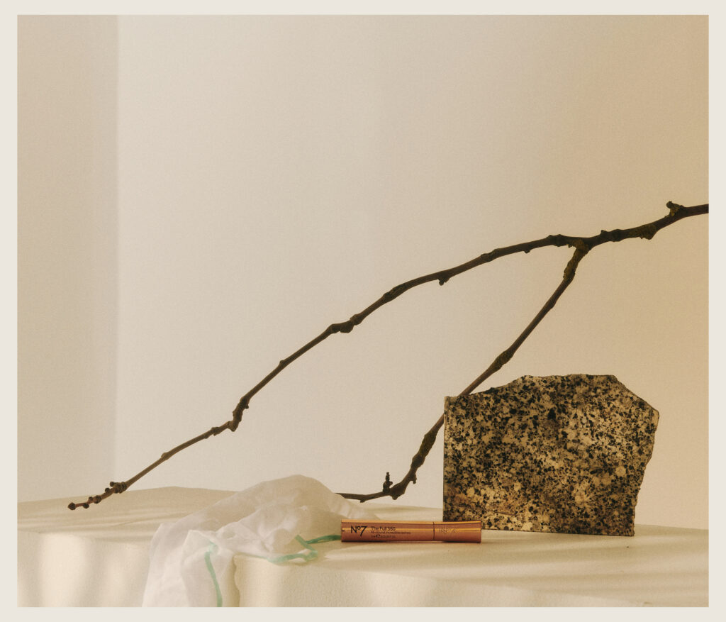 A minimal still life featuring a thin, bare branch, a speckled stone, a white cloth, and a copper-colored tube arranged on a draped, light-colored surface with a soft beige background.