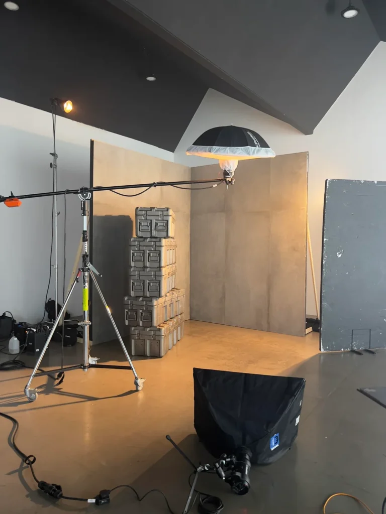 A photography studio setup with lighting equipment, tripods, a camera, and a stack of gray storage cases against a plain background wall. The space has a minimalist, industrial feel.