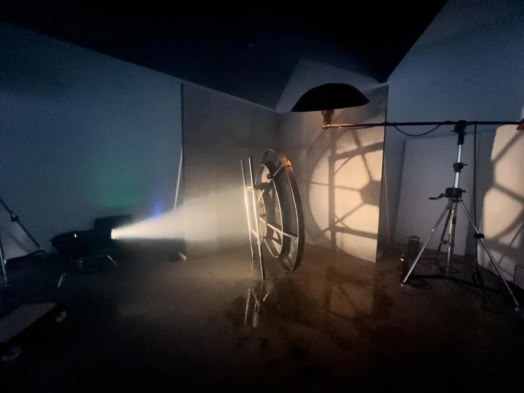 A dimly lit studio setup features a large circular object and metal bars, with dramatic shadows cast on the wall by a spotlight. Photography equipment and stands are visible on the shiny floor.